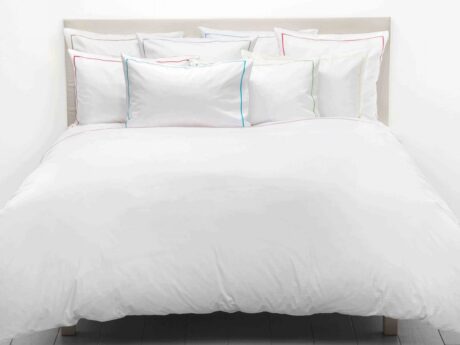 Christian Fischbacher Satin Premium bed linen styled on double bed in white Egyptian cotton satin, pillowcases shown in multiple coloured border variants including blue, red, green and grey, bright minimal bedroom