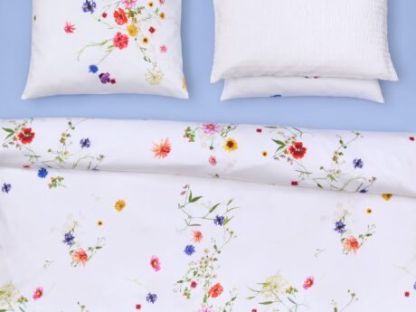 Christian Fischbacher Fleurs Volantes bed linen set styled on bed with matching pillowcases, white satin cotton with scattered wildflower pattern on blue background