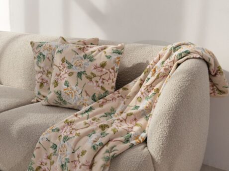 Feiler floral throw blanket and matching decorative cushions in soft beige with rose pattern displayed on a textured cream sofa
